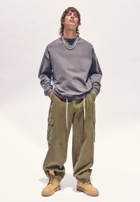 Sweatshirt gris, pantalon cargo olive et bottes beige. Le sweatshirt a une coupe décontractée, les pantalons possèdent des poches latérales et les bottes ont des lacets robustes.