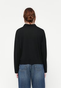 Woman with red hair in a bun wearing a black long-sleeve shirt and blue jeans, facing a plain white background.