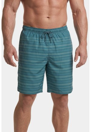Mann trägt türkisblaue Badeshorts mit horizontalen Streifen und einem Kordelzug in der Taille, steht mit entspannten Armen an den Seiten.
