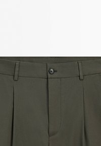 Dark green pants waist with belt loops, a front button closure, and pleated fabric details.