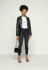 Blazer de pele sintética preta, blusa branca, jeans skinny escuros e sandálias de salto pretas. Os acessórios incluem uma pequena bolsa preta com alça de corrente.