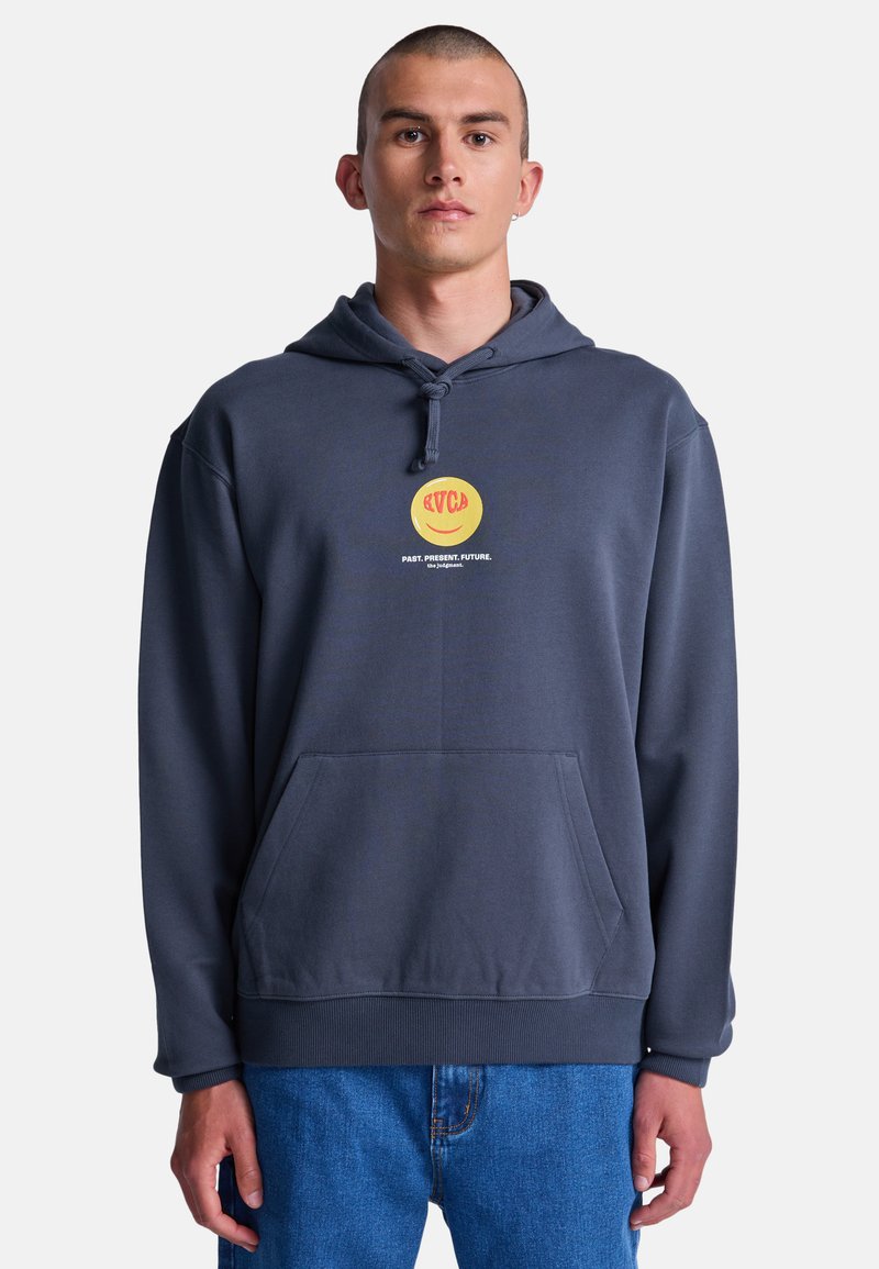 Mężczyzna w ciemnoszarej bluzie z kapturem z żółto-czerwonym logo emotikony oraz napisem „PAST. PRESENT. FUTURE.”, ubrany w niebieskie dżinsy, stojący na białym tle.