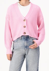Kvinna iklädd en ljusrosa grovstickad kort cardigan med stora knappar över en vit topp och högmidjade ljusblå jeans med rhinestonesmönster.