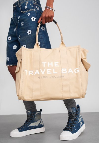 Beige kangast reisikott kahe ülemise sangaga, valge tekstiga "REISIKOTT" ja "MARC JACOBS," külgpocketitega täiendavaks salvestamiseks.