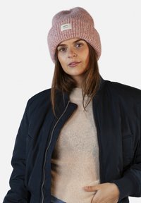 Cappello lavorato a maglia rosa con risvolto e etichetta, abbinato a una giacca bomber scura sopra un maglione chiaro. Texture morbida, design casual.