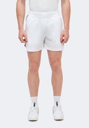 Άνδρας που φοράει άσπρα αθλητικά σορτς, άσπρες κάλτσες και άσπρα αθλητικά παπούτσια, στέκεται σε απλό φόντο.