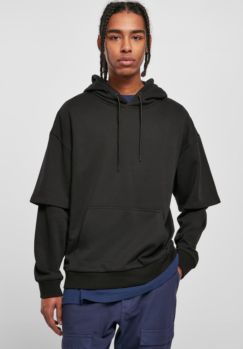 Sudadera con capucha de gran tamaño en color negro, con un bolsillo canguro, cierre con cordón y una camiseta azul de contraste debajo. Textura de tela suave.