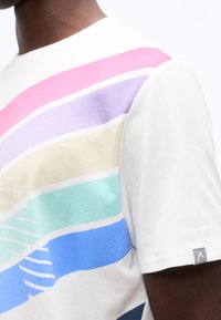 Άτομο που φοράει λευκή μπλούζα με οριζόντιες παστέλ ρίγες σε ροζ, μοβ, μπεζ, μέντα και μπλε χρώμα στο στήθος και τον ώμο.