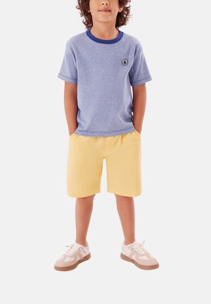 Ragazzo giovane che indossa una maglietta blu con colletto blu scuro, pantaloncini gialli e scarpe da ginnastica bianche, in piedi con le mani in tasca.