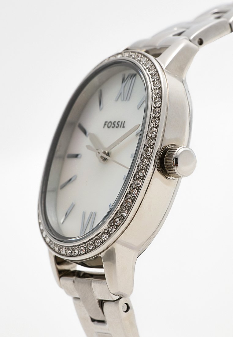 Reloj fósil plateado con un dial redondo blanco, números romanos y un bisel adornado con cristales. Pulsera de acero inoxidable con eslabones pulidos.