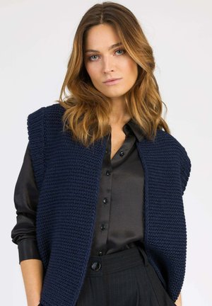 Gilet en tricot marine avec tissu texturé, emmanchures larges, porté sur une chemise noire à boutons, associé à un pantalon noir à rayures.