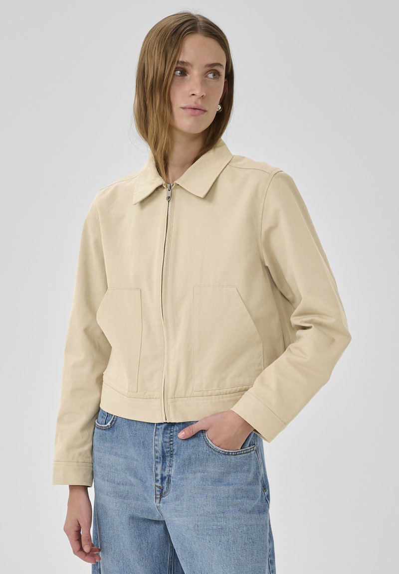 Mujer con chaqueta beige con cremallera y grandes bolsillos, y jeans azules de mezclilla, de pie frente a un fondo gris liso.