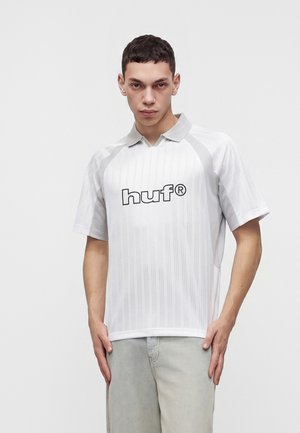 Jeune homme aux cheveux courts et foncés portant une chemise blanche à col avec manches courtes et logo "huf", associée à un pantalon de couleur claire.