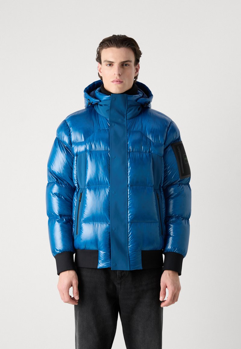 Doudoune bleue avec une finition brillante, fermeture éclair à l'avant, grande capuche et poignets côtelés noirs. Comprend une poche latérale avec un détail en métal noir.