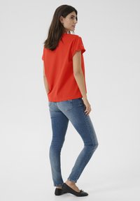 Haut rouge à manches courtes avec une coupe décontractée, associé à un jean skinny bleu. Le haut présente des accents de boutons près de l'épaule. Des ballerines noires viennent compléter le look.