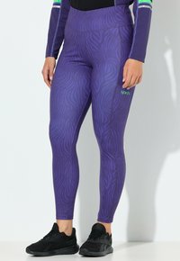 Fialové legíny se zebrovým vzorem, vyrobené z elastické látky. Mají vysoký pas a kontrastní zelené logo. Nohy obuty v černých teniskách.