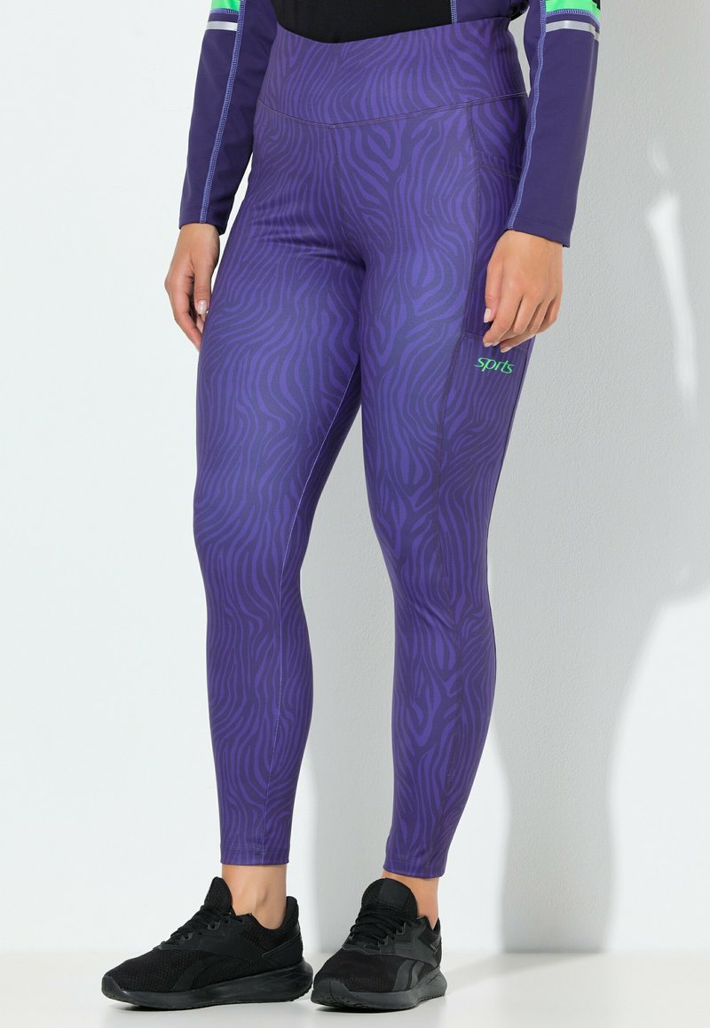 Fialové legíny se zebrovým vzorem, vyrobené z elastické látky. Mají vysoký pas a kontrastní zelené logo. Nohy obuty v černých teniskách.