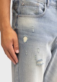 Jeans in denim blu chiaro con fitting slim, caratterizzati da strappi sfrangiati e dettagli con whiskering, design a cinque tasche e accessori in rame.