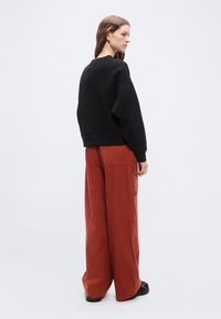 Γυναίκα με καστανά μαλλιά που φοράει μια μαύρη φαρδιά μπλούζα και φαρδιά παντελόνια σε σκουριαχτί χρώμα, στέκεται μπροστά σε ένα απλό λευκό φόντο.