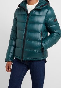 Uomo che indossa un giubbotto imbottito con cappuccio di colore teal sopra un maglione di lana bianco e jeans blu, con una mano nella tasca del giubbotto.