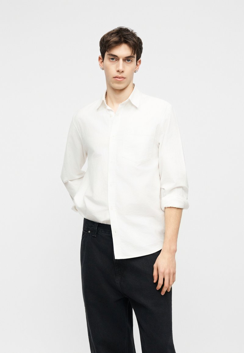 Giovane uomo con capelli corti e scuri che indossa una camicia bianca a maniche lunghe con le maniche arrotolate e pantaloni neri, su uno sfondo semplice.