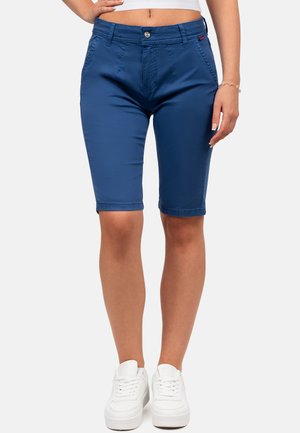 Vrouw draagt blauwe knielange shorts en witte sneakers, staand met één hand in haar zij tegen een witte achtergrond.