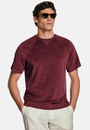 Mann, der einen dunkelroten kurzärmeligen Sweatshirt, beigefarbene Hosen und eine braune Sonnenbrille trägt, steht mit den Händen in den Taschen vor einem weißen Hintergrund.