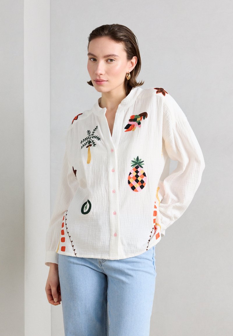Femme portant une blouse blanche boutonnée avec une broderie colorée de fruits tropicaux et de plantes, associée à un jean bleu clair, sur un fond uni.