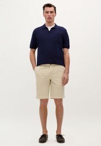 Marine polo skjorte med korte ærmer og v-hals, parret med beige shorts med opsmøg på kanten. Sorte bådsko.
