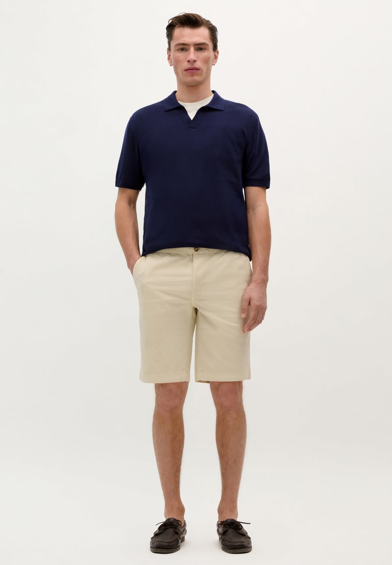 Marine polo skjorte med korte ærmer og v-hals, parret med beige shorts med opsmøg på kanten. Sorte bådsko.