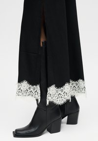 Μαύρες μπότες μέσης προς τη γάμπα με χοντρά τακούνια, φορεμένες κάτω από μαύρη φούστα με λευκή δαντέλα στο τελείωμα και μία σχισμή μπροστά.