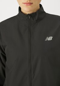 Primo piano di una persona che indossa una giacca nera New Balance con zip e colletto rialzato su uno sfondo semplice.