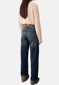 Kobieta w beżowym krótkim swetrze i szerokich, wysokich, niebieskich dżinsach z mankietami, stojąca tyłem do aparatu.