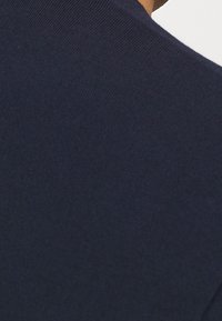 Primo piano di un tessuto blu navy che copre la spalla e la parte superiore della schiena di una persona, con una scollatura visibile e un tono della pelle nell'angolo in alto a destra.