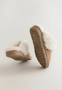 Tummanruskeat mokkasandaalit, joissa on valkoinen tekokarva vuori, pehmeä pinta ja kumiset pohjat, joissa on teksturoitu pito. Alapuolelta näkyy pohjan kuvio.