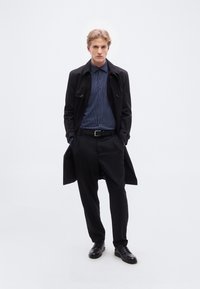 Μαύρο παλτό τρεντσόρ πάνω από μπλε πουκάμισο με πουά, συνδυασμένο με μαύρο παντελόνι και παπούτσια. Λείος υφής, επίσημο σχέδιο, με λεπτομέρειες όπως κουμπιά και ζώνη.