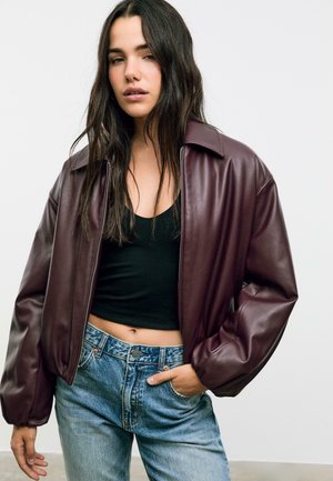 Veste en faux cuir bordeaux avec un large col et des poignets côtelés, portée sur un crop top noir et associée à un jean bleu clair.