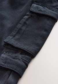 Tissu bleu foncé avec une texture lisse, présentant un design de manchette avec bords repliés et des détails de couture visibles. Deux poches inclinées près de la manchette.