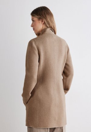 Kvinde set fra siden iført en beige lang uldfrakke med høj krave og hænderne i lommerne mod en ensfarvet lys baggrund.