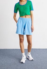 Zelený zkrácený zipový svetr s modrými akcenty, spárovaný se světle modrými šortkami s elastickým pasem. Modelka má na sobě bílé tenisky s modrými detaily.