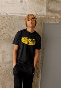 Sort bomulds T-shirt med gul grafisk tryk, der viser stiliseret tekst; parret med sorte sportbukser; står op ad en struktureret betonvæg.