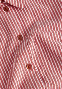 Czerwona i biała koszula zapinana na guziki, z kieszenią na piersi i brązowymi guzikami. Tkanina ma teksturowaną powierzchnię, z równomiernie rozmieszczonymi pionowymi paskami.