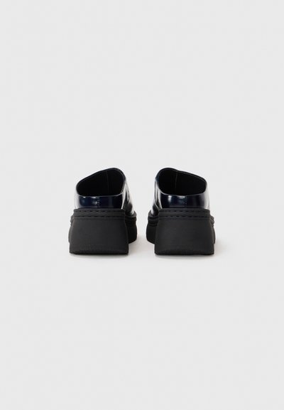 Une paire de chaussures noires en cuir à enfiler avec des semelles épaisses, vue de dos sur fond blanc.