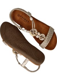 Des sandales plates argentées avec des brides tressées, des accents décoratifs en perles et une semelle marron texturée avec des motifs en vagues. Logo de la marque visible à l'intérieur.