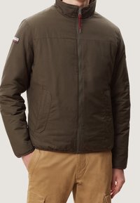 Brun isoleret jakke med høj krave, frontlynlås og to sidelommer. Har et logo-patch på venstre skulder.