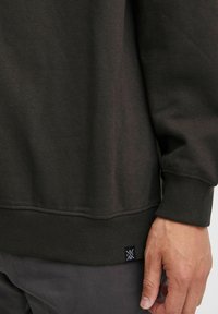 Mørkegrøn sweatshirt med ribbede manchetter og kant; glat tekstur med et lille mærke nederst. Nærbillede af stofdetaljer.