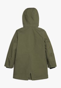 Parkas oliwkowozielona z kapturem, długimi rękawami, prostym krojem oraz rozcięciem z tyłu dla wygody ruchu. Charakteryzuje się widocznymi przeszyciami i prostym wzornictwem.