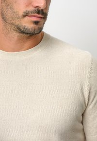 Hekelcreme Pullover mit strukturierter Oberfläche, rundem Halsausschnitt und dezenten Nahtdetails. Der Stoff wirkt weich, leicht und atmungsaktiv.