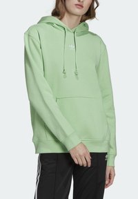 Ljusgrön hoodie med kängurufickor, dragsko på huva och vit logotyp. Mjukt bomullsmaterial med avslappnad passform och ribbade ärmslut.