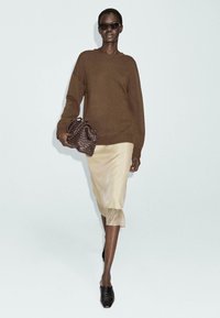 Donna che cammina in un maglione marrone, gonna di seta beige, scarpe nere, mentre tiene in mano una borsa a tracolla intrecciata marrone e indossa occhiali da sole scuri su uno sfondo neutro.